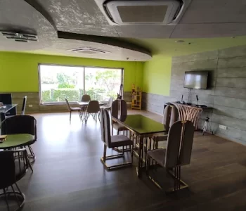 Bright lounge with lime-green walls, wooden tables and upholstered chairs, large windows at the far end letting in natural light, and a gray wood-paneled wall with a TV and cabinet nearby.